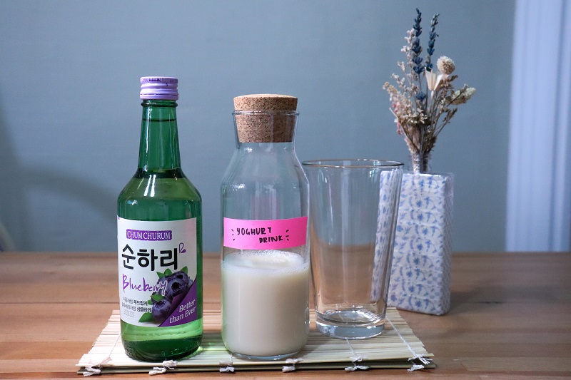 A bottle of blueberry soju, yoghurt drink and glass displayed on a table.